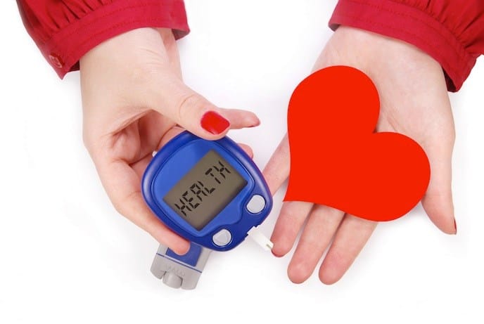 Two female hands with the left holding a blood glucose meter and the right holding a red cardboard heart.