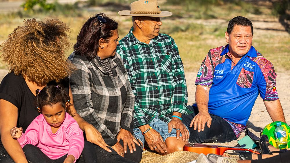 A group of Aboriginal and Torres Strait Island people are sitting in the Australian bush looking towards a Diabetes Australia First Nations health worker while talking.