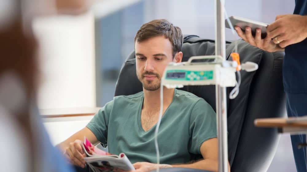A young man sits in a hospital chair reading a magazine with drips and an iv in the frame.
