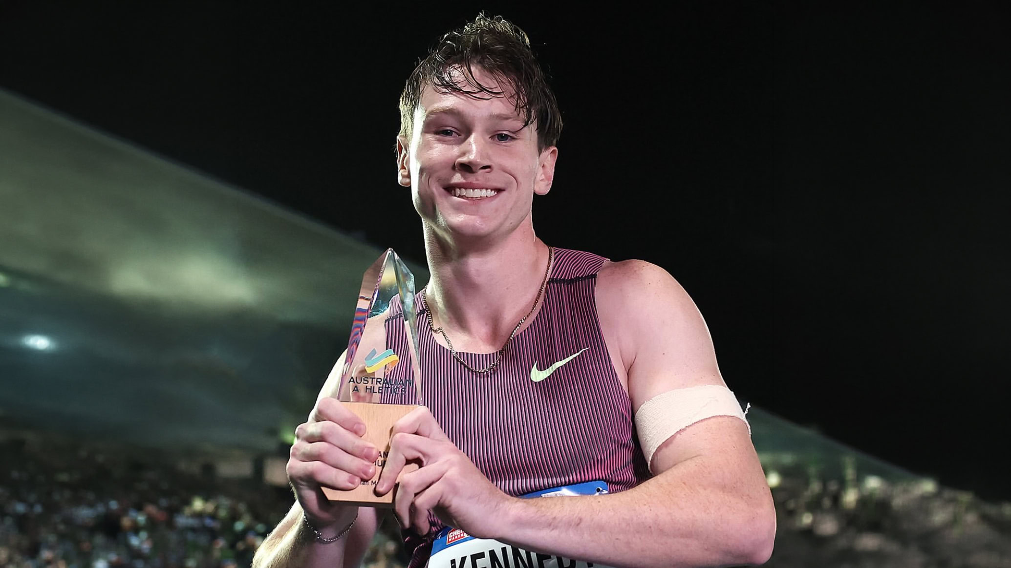 Lachie looks down at the camera while holding the Australian Athletics 100m Champion trophy.