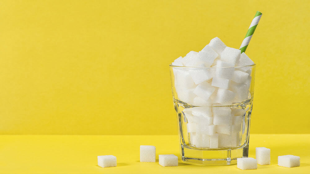 Against a bright yellow background, a glass with white sugar cubes has a blue and white straw sticking out of it, with sugar cubes lying around its base.