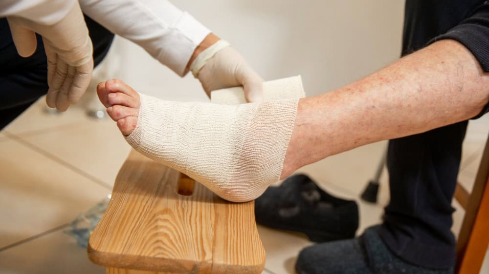 A foot propped on a wooden stool is carefully bandaged by a health professional wearing gloves.