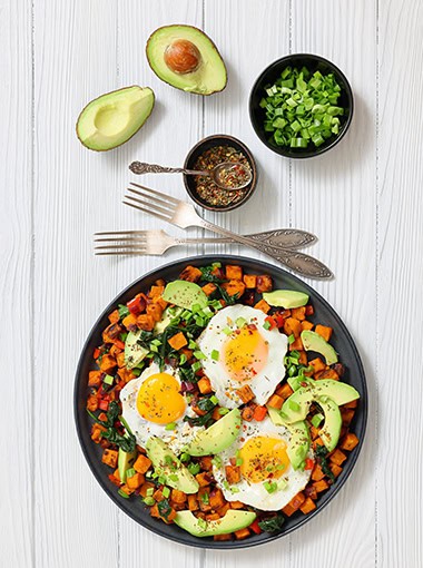 Sweet potato cubes served with sunny-side-up eggs and fresh avocado slices on a plate on white wooden table with seasoning and forks