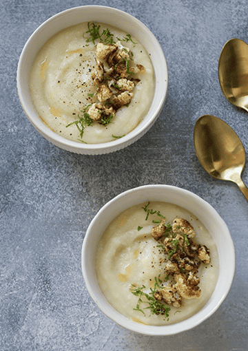 Two bowls of creamy cauliflower soup with a cauliflower garnish, accompanied by two gold spoons